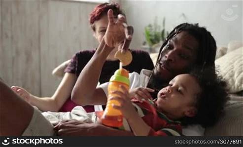 Loving interracial parents trying to convince their curly mixed race toddler son to drink juice from feeding bottle at home. Affectionate african american father with dreadlocks fooling around, trying to give a drink to his little kid. Slow motion.