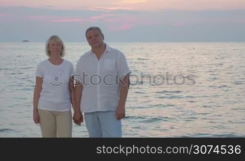 Loving mature couple standing on the beach. At first they holding hands and they embracing each other. Sea and evening sky in background