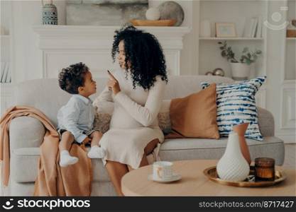 Loving mom young pregnant woman sitting on sofa in living room, communicating with cute little son with curly hair, touching his nose with finger, boy listening with concentration. Family concept. Portrait of loving afro american pregnant mother explaining little son about her pregnancy