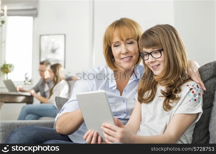 Loving mother and daughter using tablet PC with family sitting in background at home