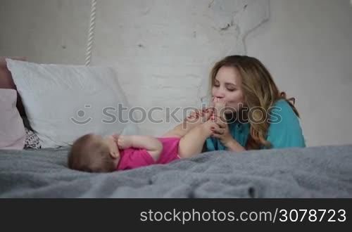 Loving mother kissing infant child feet symbolizing tenderness and care. Beautiful woman expressing her love, bonding with her baby girl in bedroom. Slow motion. Steadicam stabilized shot.
