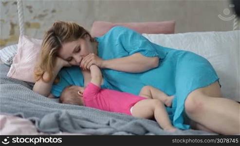 Loving mother lying on her side on the bed and breastfeeding infant child in modern styled bedroom. Beautiful affectionate mother kissing baby arm with love while feeding breast to her baby girl at home.