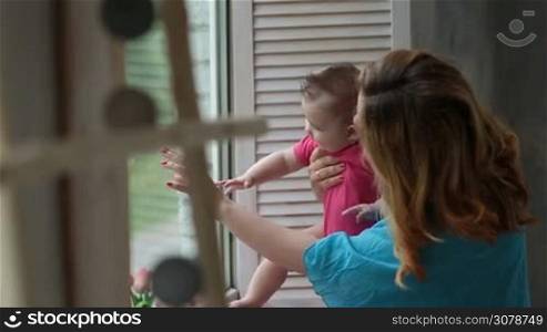 Loving mother standing next to window, holding her cute infant child, looking out of the window and telling kid about nature outside. Adorable baby girl discovering the world while looking out of the window together with her beautiful mom.