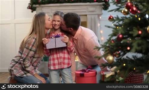 Loving parents kissing happy daughter in Christmas decorated room. Adorable excited little girl holding xmas gift box and affectionate parents kissing her cheeks during winter holidays. Dolly shot.