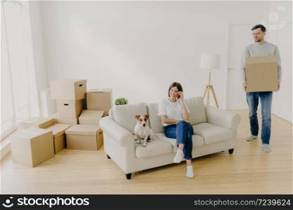 Loving spouses pose in rented apartment, woman relaxes on couch with pet, speaks via modern smartphone, smiles happily, busy husband holds cardboard box with belongings, justed moved in new apartment