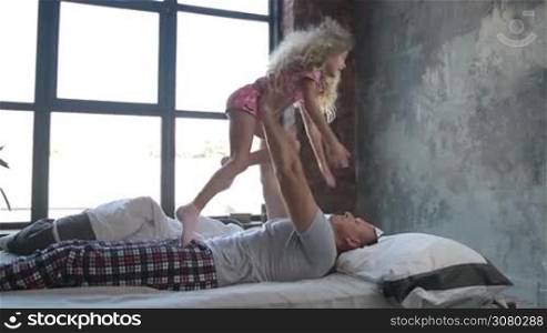 Loving young dad lying on his back, lifting his cute curly daughter up over his head letting child fly in grunge styled bedroom. Caring handsome father and adorable little girl having fun in bed while spending free time together. Slo mo. Dolly shot.