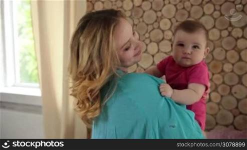 Loving young mother holding and hugging her cute infant child, slowly dancing in modern living room. Happy family enjoying time together at home. Slow motion. Steadicam stabilized shot.