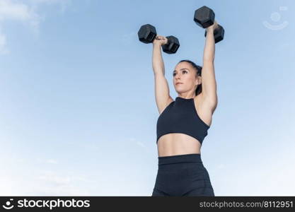 Low ang≤of strong fema≤ath≤te in sportswear holding heavy dumbbells in raised arms against blue sky during∫ense fit≠ss workout in daytime. Sportswoman lifting dumbbells against blue sky