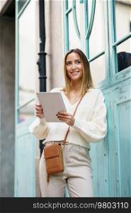 Low angle of cheerful woman in stylish wear with brown shoulder bag standing on city street while browsing tablet. Positive female using tablet on town street