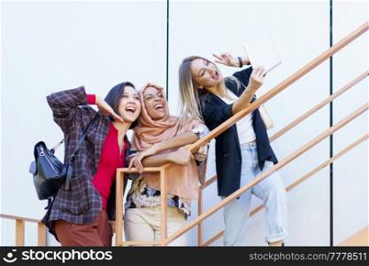 Low angle of delighted young multiracial female students in trendy outfit gesturing and smiling while taking selfie on tablet standing on stairs on city street. Excited young multiethnic female millennials smiling and taking selfie on tablet