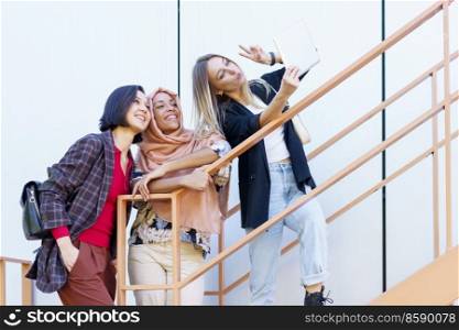 Low angle of delighted young multiracial female students in trendy outfit gesturing and smiling while taking selfie on tablet standing on stairs on city street. Excited young multiethnic female millennials smiling and taking selfie on tablet