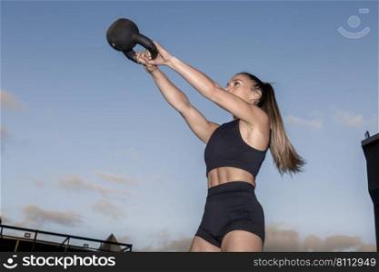 Low angle of determined female athlete in activewear with ponytail lifting heavy kettlebell in outstretched arms during fitness workout on sports ground in evening. Sportswoman exercising with kettlebell on sports ground
