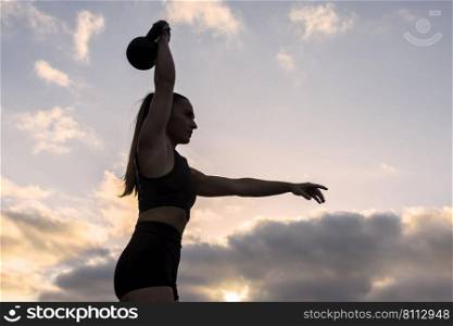 Low angle of fit female athlete in sportswear with ponytail raising arm with heavy kettlebell over head while exercising against cloudy sundown sky. Strong sportswoman lifting kettlebell at sunrise