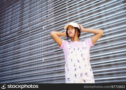 Low angle of glad Asian female in casual clothes, wearing cap, looking away standing near modern building.. Low angle of glad Asian female in casual clothes, wearing cap, standing near modern building.