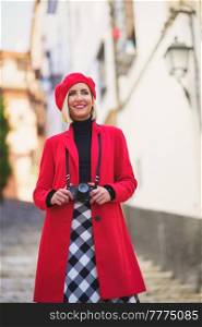 Low angle of happy young lady with blond hair, in stylish warm clothes and beret, smiling and looking away while admiring and photographing town on camera. Smiling female tourist with photo camera standing on stairs on city street