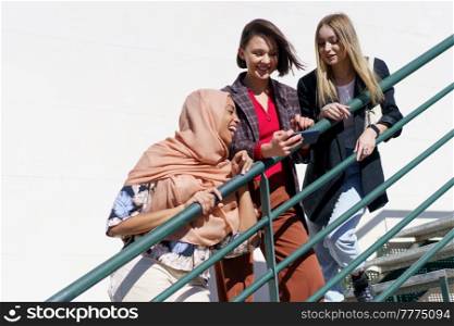 Low angle of joyful young Muslim woman in hijab, laughing while watching photo on smartphone with happy female friends, standing together on metal staircase on sunny day. Diverse cheerful female students smiling while sharing smartphone on staircase