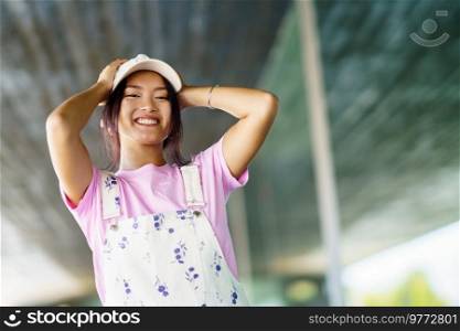 Low angle of positive young Asian female looking at camera smiling with happiness. Chinese woman in casual clothes and cap, near a modern building.. Positive young Asian female looking at camera smiling with happiness near a modern building