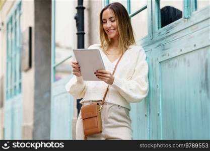 Low angle of young smiling female in stylish beige outfit with bag leaning on wall while using tablet outside. Cheerful woman with tablet standing on street