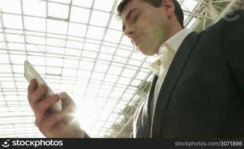 Low angle shot of a young businessman using smart phone in trade center or office builidng. Metal and glass roof with bright sun flare in background