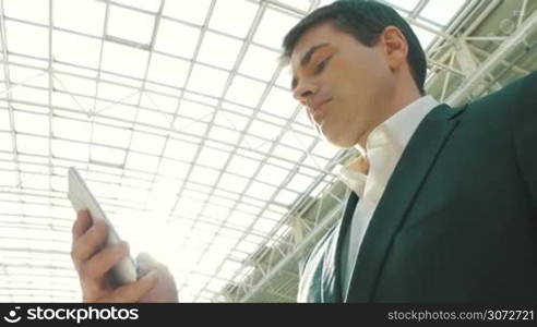 Low-angle shot of a young businessman using smartphone in modern building with glass roof. He&acute;s brightly lit with sunlight.