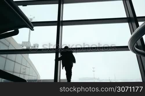 Low angle shot of child coming up to the big window and looking out of it while woman with trolley bag passing by