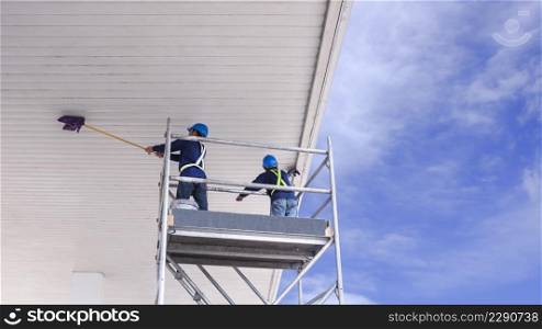 Low angle view of 2 cleaner workers on scaffolding using flat mops to cleaning white ceiling roof of petrol station