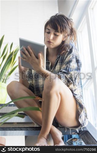 Low angle view of a beautiful sensual woman wearing shirt with ponytail sitting on staircase at home while holding a tablet pc next to a window in a sunny day. Isabella Antonelli
