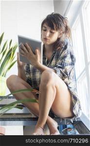 Low angle view of a beautiful sensual woman wearing shirt with ponytail sitting on staircase at home while holding a tablet pc next to a window in a sunny day. Isabella Antonelli