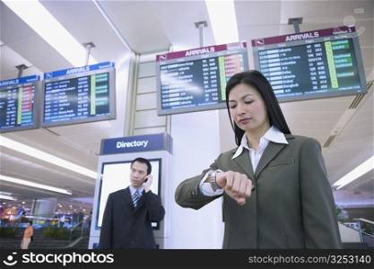 Low angle view of a businesswoman looking at her wristwatch with a businessman talking on a mobile phone behind her at an airport