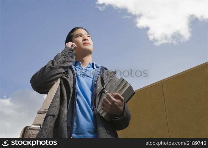Low angle view of a young man holding books and talking on a mobile phone