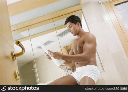Low angle view of a young man sitting in the bathroom reading a newspaper