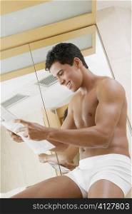 Low angle view of a young man sitting in the bathroom reading a newspaper