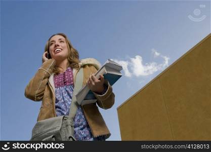Low angle view of a young woman holding books and talking on a mobile phone