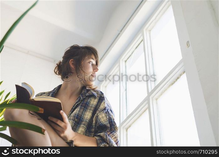 Low angle view of beautiful sensual woman wearing shirt with ponytail sitting on staircase at home while holding a book next to a window in a sunny day. Rafa Fernandez