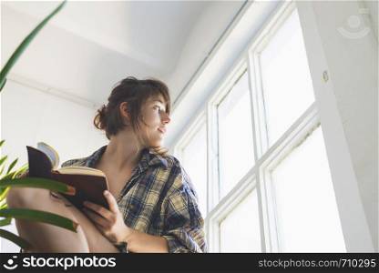 Low angle view of beautiful sensual woman wearing shirt with ponytail sitting on staircase at home while holding a book next to a window in a sunny day. Rafa Fernandez
