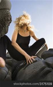 Low angle view of Caucasian mid-adult woman crouching on rocks with hair blowing wildly in wind.