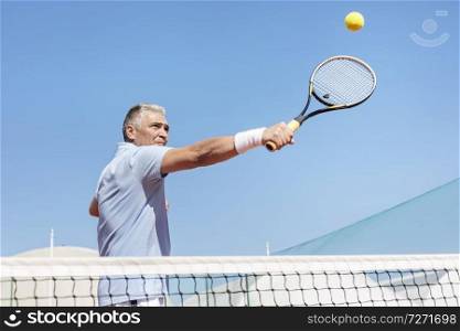 Low angle view of confident mature man hitting tennis ball with racket on court against clear blue sky