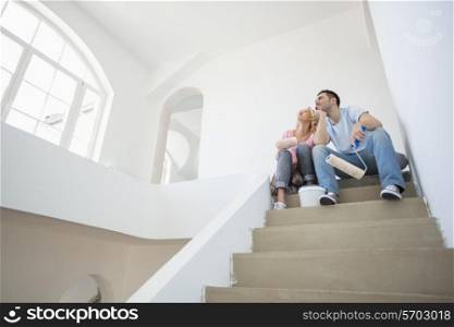 Low angle view of couple with painting tools sitting on steps in new house