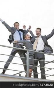 Low angle view of excited businesspeople with arms raised standing on terrace against clear sky