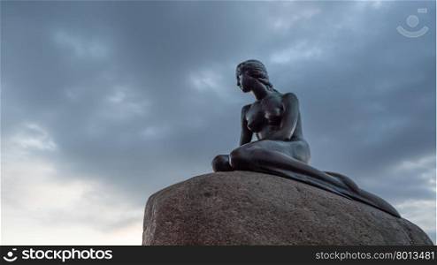 Low angle view of Little Mermaid statue on large boulder looking away in Denmark under cloudy sky. Low angle view of Little Mermaid statue in Denmark
