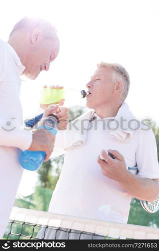 Low angle view of thirsty men drinking while standing by senior friend on tennis court against clear sky