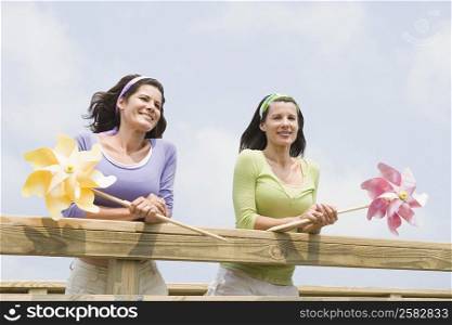 Low angle view of two mature women leaning on a wooden fence and holding pinwheels