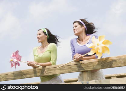 Low angle view of two mature women leaning on a wooden fence and holding pinwheels