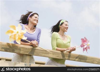 Low angle view of two mature women leaning on a wooden fence and holding pinwheels