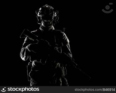 Low key studio portrait of army special forces elite soldier with hidden behind mask and glasses face, battle helmet, tactical radio headset, standing with assault rifle equipped silencer in darkness. Army special forces soldier low key studio shoot