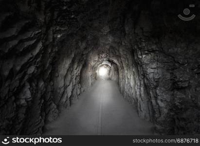Low light picture with a road passing through a tunnel built in a mountain, with stone walls and a bright light at its end. Tunnel located in the Swiss Alps.