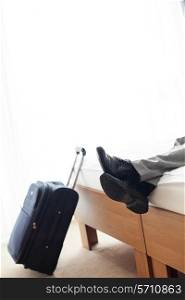 Low section of businessman lying on bed beside luggage in hotel room