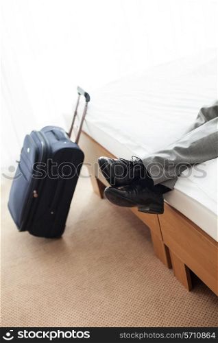 Low section of businessman lying on bed with luggage on floor in hotel room