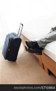 Low section of businessman lying on bed with luggage on floor in hotel room
