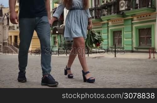 Low section of loving couple holding hands walking along city street during romantic date. Closeup of male and female legs in casual footwear going down the street while spending leisure together outdoors. Slow motion. Steadicam stabilized shot.